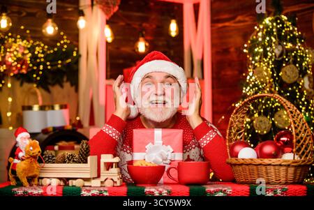 Joyful grandpa with gift. Dreamy grandfather in Santa hat. Christmas eve. New year. Cozy home. Hot beverage and cookies. Joyful atmosphere. Senior Stock Photo