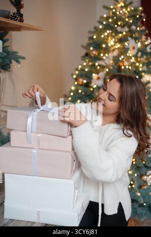 Woman in white sweater holding stack of wrapped gifts near Christmas tree. Stock Photo