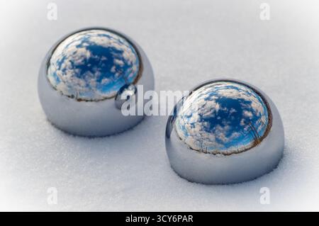 Two metallic spheres placed on snow reflect a vibrant sky with scattered clouds. Stock Photo