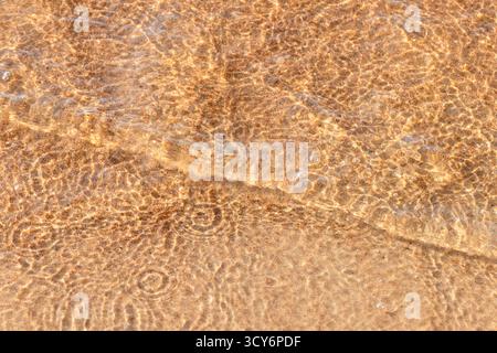 A close view of shallow, sunlit water over wet sand creating intricate ripple patterns and gentle waves. Warm golden tones evoke calm seaside moments, Stock Photo