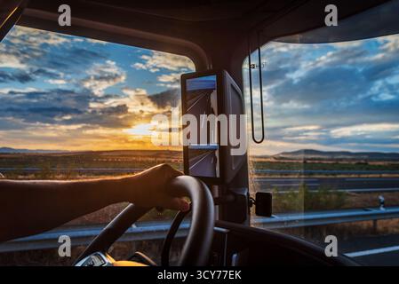 Truck cabin view with driver’s hand on the wheel under a colorful sunset sky Stock Photo