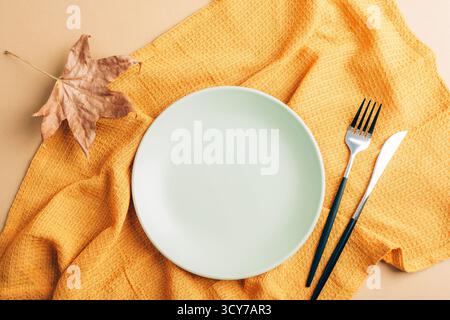 Empty green plate with fork and knife and autumn leaf on yellow napkin, top view. Minimal tableware and dining background concept. Flat lay, mockup. Stock Photo