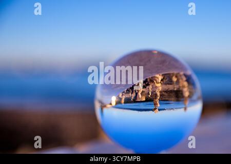 Glass ball on a seaside wall producing an inverted view of stacked stones and water in Lisbon, Portugal, photographed at golden hour with shallow DOF. Stock Photo