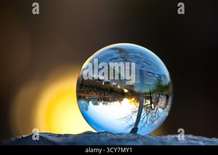 Crystal ball on a stone ledge reflecting an inverted riverside street and setting sun glow, captured with shallow depth of field at golden hour light. Stock Photo