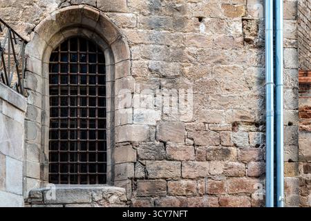 Arched window with a massive metal grille against the backdrop of an old stone wall with drainpipes. From the "Windows of the World" series. Stock Photo