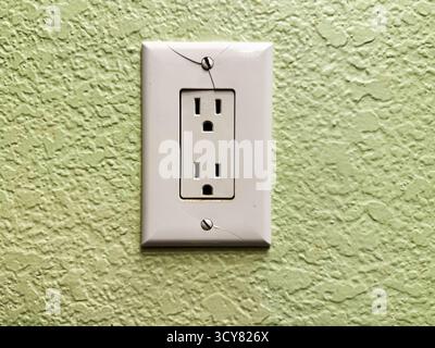 Cracked wall electrical outlet with damaged plastic faceplate on textured green interior wall, home maintenance and electrical safety concept Stock Photo