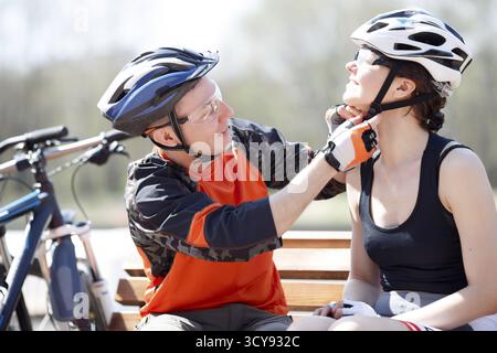 Guy, girl sitting on bench, putting on helmet in park Stock Photo