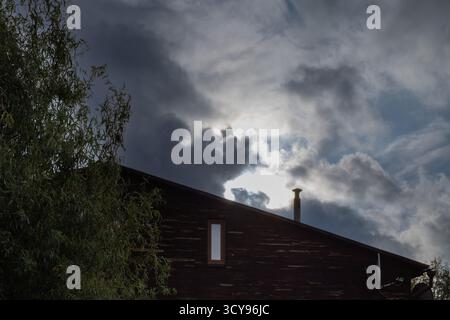 Dark wooden house with small window and chimney against cloudy sky and sunlight. Autumn rural landscape. Stock Photo