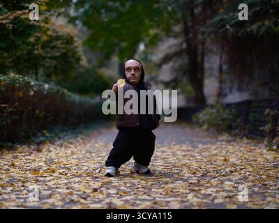 Curious man with dwarfism standing on park path covered with yellow autumn leaves, holding single yellow leaf. Unique individual interacting with natu Stock Photo