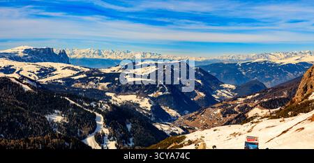 Expansive winter panorama showing a vast Dolomites valley landscape with snowy mountains, dark forests, and a sunlit village below a bright blue sky. Stock Photo