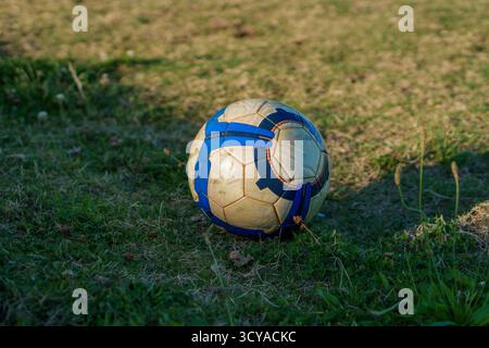 An old, dirty soccer ball with blue, white, and orange patterns sits on a patch of green and dry grass. Stock Photo