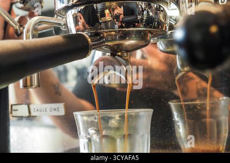 Close-up of espresso pouring into glass cups from a chrome coffee machine at Cafeteria Triple Five, with barista reflection visible. Stock Photo