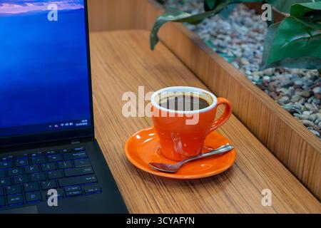 A cup of black coffee in an orange mug placed on a wooden table beside an open laptop, symbolizing remote work, productivity and modern workplace life Stock Photo