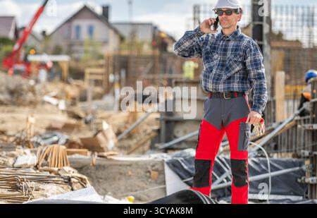 A construction worker in a plaid shirt and hard hat stands near building materials while talking on his phone. Stock Photo