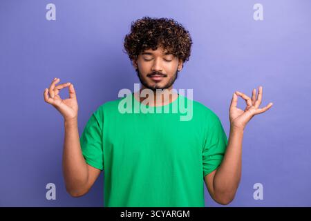 Relaxed young man in a green t-shirt meditating with a calm expression and hand gesture against a violet background Stock Photo