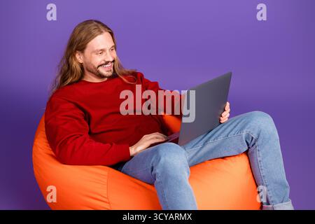 Young man with long blond hair in a casual red sweater sits on an orange bean bag and uses a laptop against a purple background Stock Photo