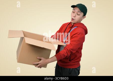 young man in red hoodie carrying open cardboard box, surprised expression, indoor studio photography shot Stock Photo