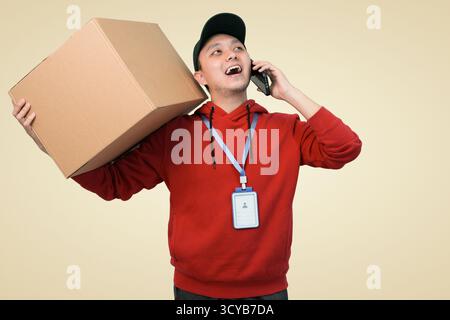 Happy delivery package worker wearing red hoodie balances box while talking on phone Stock Photo