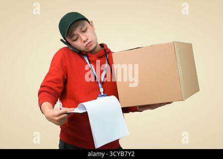 Busy delivery man on phone with large cardboard box and paper roll in casual red hoodie Stock Photo
