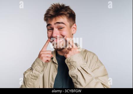 A young man wearing a light-colored shirt smiles playfully, using his fingers to point at his teeth. The neutral backdrop highlights his joyful expres Stock Photo