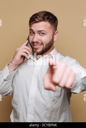 A young man with a beard smiles confidently while talking on the phone. He points directly at the camera, creating a lively and engaging scene in a si Stock Photo