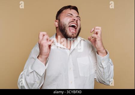 A man shows intense frustration with his hands clenched, mouth open in a scream. He is dressed in a white shirt against a simple beige background, emp Stock Photo