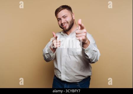 A smiling man stands confidently in a studio. He is wearing a white shirt and blue jeans, pointing playfully with both hands toward the camera. His ch Stock Photo