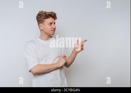 A young man stands confidently, pointing to his right with a smile. He is dressed in a simple white t-shirt and has styled blonde hair, creating a rel Stock Photo