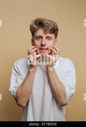 A young man stands in a studio against a plain background, displaying fear through his wide eyes, open mouth, and hands on his face. His expression is Stock Photo