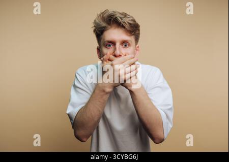 A young man stands against a plain background, covering his mouth with both hands while expressing surprise or shock. His wide eyes reveal strong emot Stock Photo