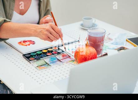 During creative hobby session, the woman is focused on painting pomegranate fruit in her sketchpad with brush and palette. close-up shot highlights th Stock Photo