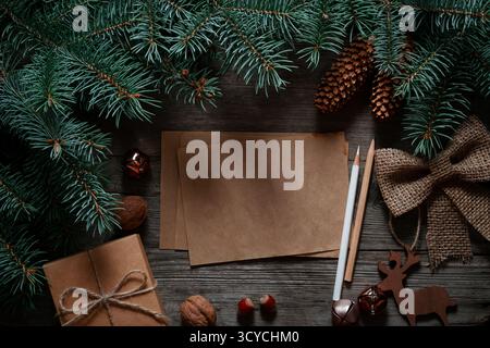 A blank kraft paper card is the center of a dark, rustic Christmas flat lay with fir branches, a gift, and decorations. Stock Photo