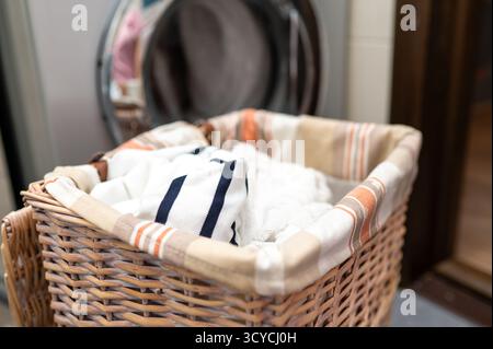 Wicker laundry basket filled with dirty clothes placed in front of washing machine, concept of household chores and home cleaning Stock Photo