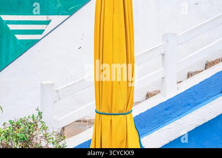 Vibrant abstract image focusing on a folded yellow umbrella pole against a wall with staircase and strong blue, white, and green geometric lines Stock Photo