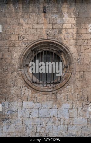 Close-up of a circular stone-framed window set in a weathered brick wall, secured with old, rusty iron protective bars Stock Photo