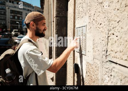 Young man with backpack pressing a door intercom button on a sunny street. Concept of arrival, communication, and urban living. Stock Photo