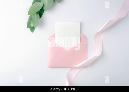 Pink envelope with blank card and ribbon on white background. Holiday greeting card, invitation, anniversary concept. Top view, flat lay, mockup. Stock Photo
