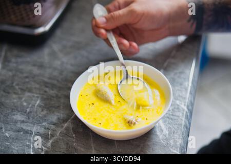 Close-up of Tattooed Hand Scooping Creamy Yellow Chicken Soup with Spoon from White Bowl on Gray Marble Kitchen Countertop Stock Photo