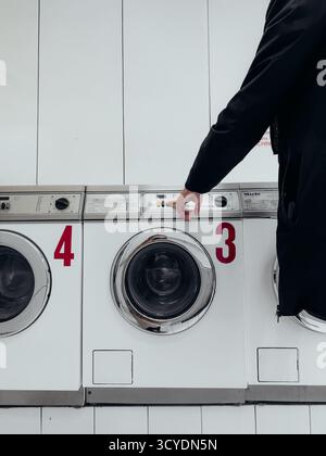 Man in black jacket pressing button on industrial washing machine number three in modern laundromat clean white minimal interior gesture control daily Stock Photo