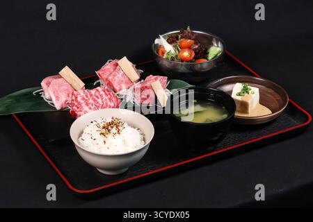 Wagyu Beef Yakiniku Bento Set on a black tray include raw Wagyu beef slices, rice, miso soup, tofu, and salad isolated on dark background side view of Stock Photo