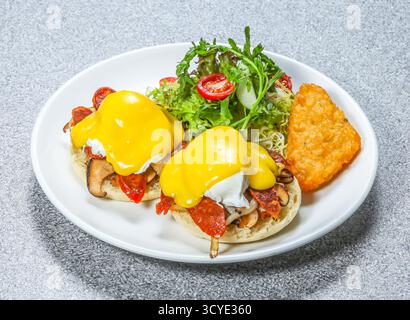 Eggs Benedict with two poached eggs with hollandaise sauce on English muffins, with salad and a hash brown served on a wooden board side view of hong Stock Photo