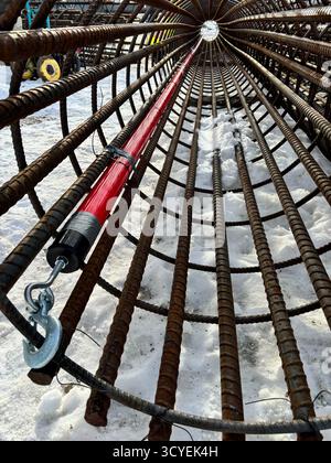 Close-up inside view of a cylindrical rebar reinforcement cage with red conduit and lifting hook, used for concrete foundation construction in winter. Stock Photo