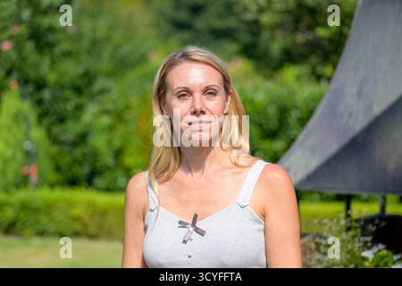 A blonde woman in a light gray top poses outdoors in a garden setting, looking confidently into the camera. Stock Photo