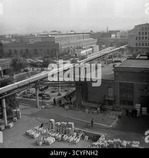 This archival 1970s B&W photo gives a high-angle panoramic view of the Slavyansk Armature-Insulator Plant (AIZ) in Sloviansk, USSR. The shot captures the vast industrial yard, dominated by a large network of elevated, insulated pipelines on concrete supports. In the foreground, numerous wooden crates and piles of finished products (insulators, fittings) are stored. Workers are walking in the yard, and large factory workshops are visible in the background. This is a document of Soviet industrial infrastructure Stock Photo