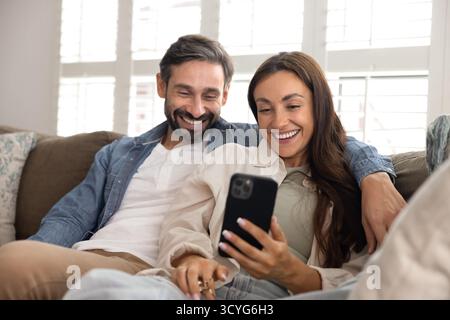 Smiling wife resting on cozy couch with husband using phone Stock Photo