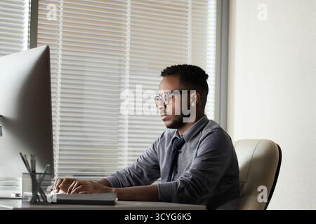 Young handsome black manager working late in office, sitting at computer and corresponding with business partners, side view Stock Photo