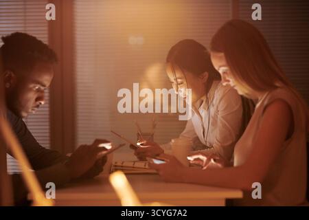 Multiethnic team of young office workers sitting at table in dark break room, resting and checking social networks on their gadgets Stock Photo