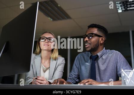 Colleagues working together at night. Young Caucasian woman and African American man sitting at table in office and looking at computer screen, low angle view Stock Photo