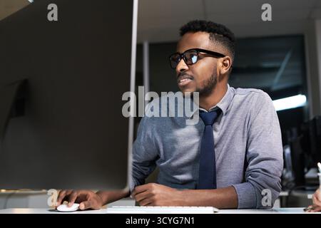 Young handsome black manager in glasses sitting at office table and working on computer till late night, low angle view Stock Photo