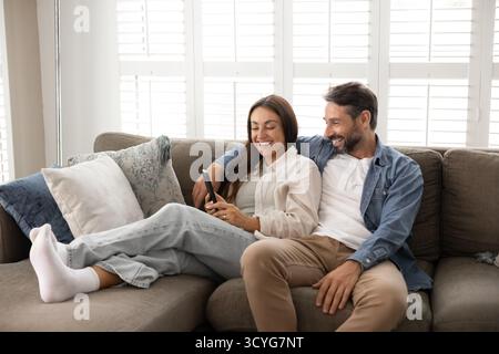 Happy family couple hugging on sofa at home using smartphone Stock Photo